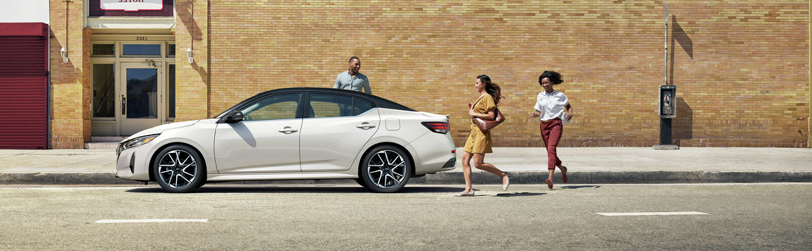 White Nissan Sentra sedan parked on a city street with pedestrians