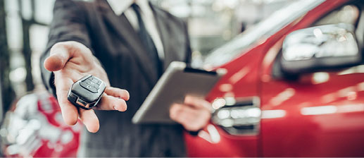 Auto dealership sales representative holding out a vehicle key fob beside a red SUV
