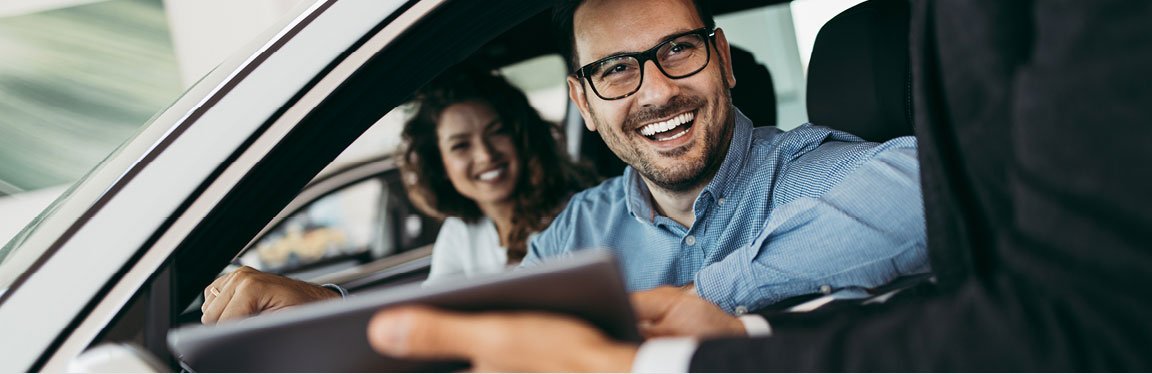 Smiling couple completing a vehicle purchase with a dealership representative while seated inside their new car