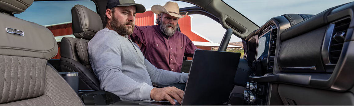 Two men using a laptop inside a Ford F-150 pickup truck near a rural farm